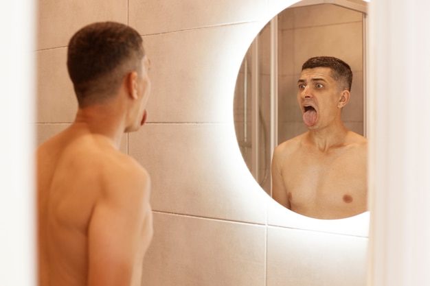 욕실 거울 앞에서 혀를 내미는 벌거벗은 상체를 가진 젊은 성인 갈색 머리 남성의 실내 사진, 아침에 위생 절차를 수행합니다.