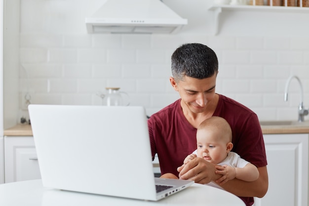 Indoor shot of young adult father wearing maroon casual t shirt sitting with little daughter or son in front of notebook, looking at baby with great love, freelancer work while taking care of child.