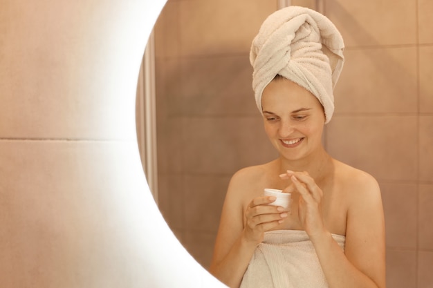 Indoor shot of young adult female taking care of her skin, posing in bathroom in front of mirror with cosmetic cream in hands, standing with bare shoulders and towel on head.