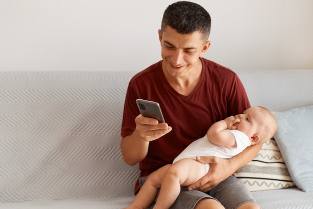 Indoor shot of young adult male wearing maroon casual style t shirt, sitting on gray sofa in room, holding infant baby, using smart phone, smiling happily.