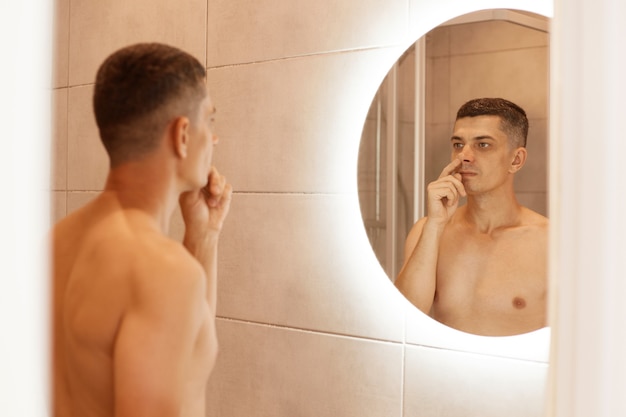 Indoor shot of young adult naked brunette male picking his nose in bathroom, standing with bare body, reflection in the mirror of man after taking shower.