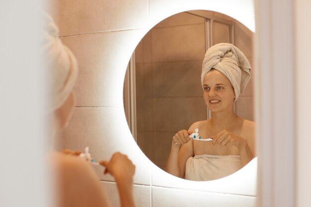 Indoor shot of young adult woman brushing teeth in bathroom, looking at her reflection in the mirror, standing with bare shoulders and white towel on her hair.