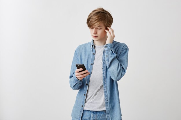 공정한 머리를 가진 젊은 백인 남자의 실내 샷 대학에서 수업 후 휴식 데님 셔츠를 입고. 스마트 폰의 음악 앱을 사용하여 흰색 이어폰으로 음악을 듣는 사람.