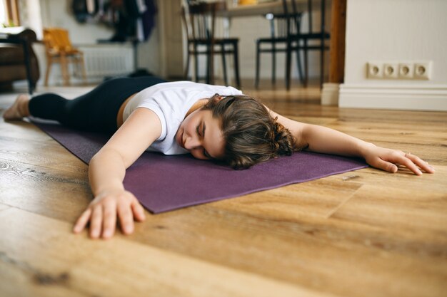 Indoor shot of young Caucasian woman in sportswear lying on stomach on mat keeping arms stretched out to front, having rest in calming pose between asanas, doing hatha yoga at home, relaxing body