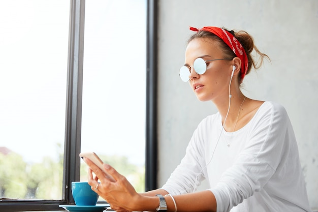 Indoor shot of young cute woman wears red headband and sunglasses, listens favourite composition from playlist via mobile phone, connected to wireless internet and earphones at cozy coffee shop