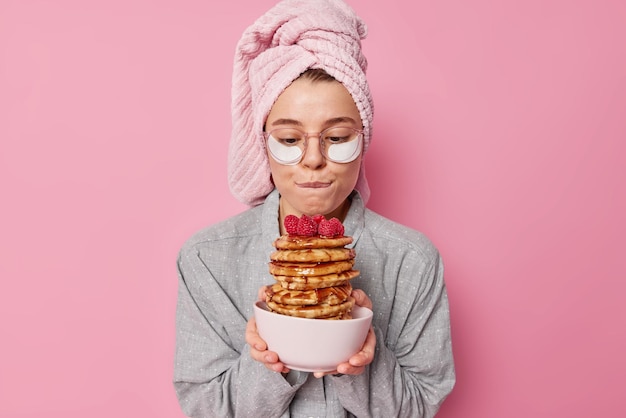 Foto gratuita l'inquadratura in interni di una giovane modella che morde le labbra guarda appetitose frittelle con sciroppo e lamponi preparate una deliziosa colazione indossa un asciugamano avvolto in pigiama sulla testa occhiali applica cuscinetti di bellezza