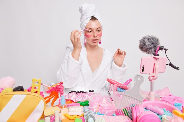 Indoor shot of young female model films make up tutorial video applies eye shadow and pink beauty patches under eyes gives online visage classes 