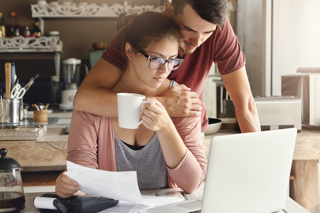 財政的ストレスに直面している若い不幸な白人家族の屋内撮影。眼鏡をかけて美しい女性が後ろに立って抱擁している夫と書類をやりながらお茶を飲む