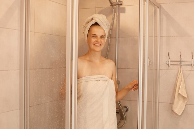 젊은 여성의 실내 사진이 샤워실에서 나오며 하얀 수건에 싸여 서서 행복한 표정으로 카메라를 바라보며 아침 일과를 하고 있습니다.
