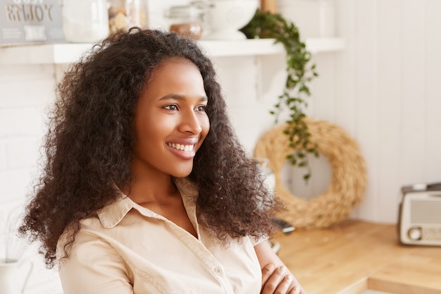 Indoor side view of amazing happy young African American female with Afro hairstyle smiling broadly, keeping arms on her chest, listening to nice music on radio, baking pie in cozy kitchen