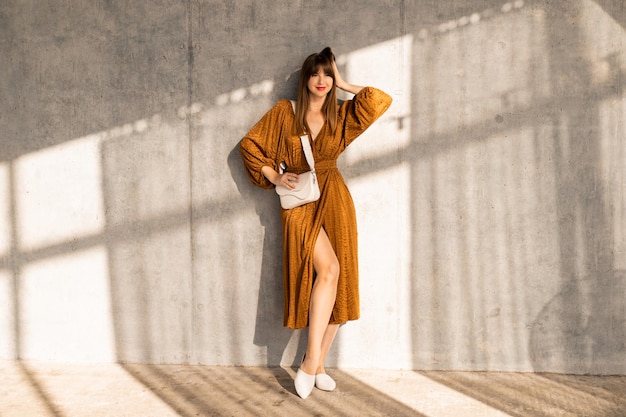 Indoor studio photo of elegant brunette woman in stylish dress standing over urban concrete wall.