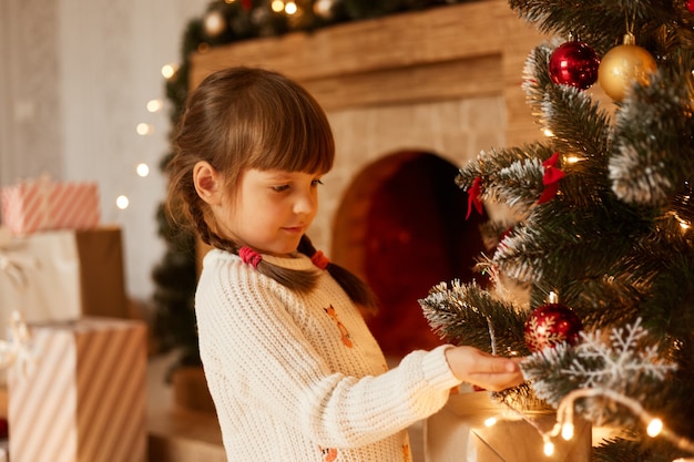 Indoor studio shot of charming girl wearing white sweater and having pigtails, decorating Christmas tree, standing near fireplace, having festive mood.