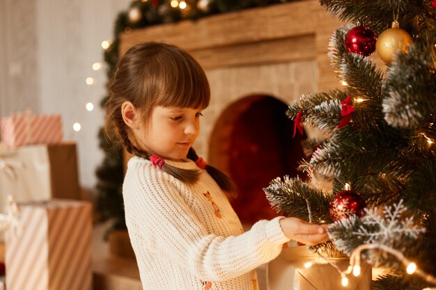 Крытый студийный снимок очаровательной девушки в белом свитере с косичками, украшающей елку, стоящей у камина и праздничного настроения.