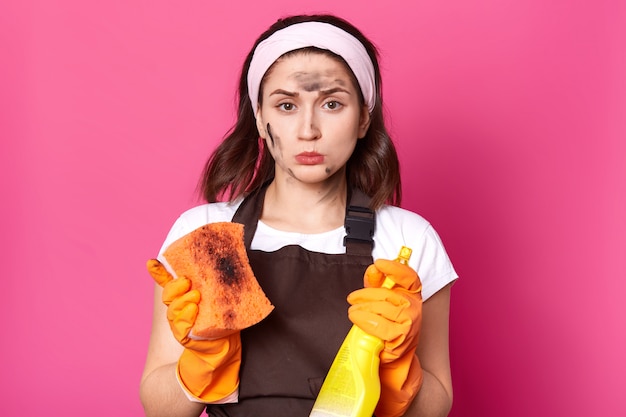 Free photo indoor studio shot of upset disappointed housewife looking directly at camera, holding dirty washcloth and detergent, wearing cleaning gloves, headband and t shirt, brown apron. household concept.