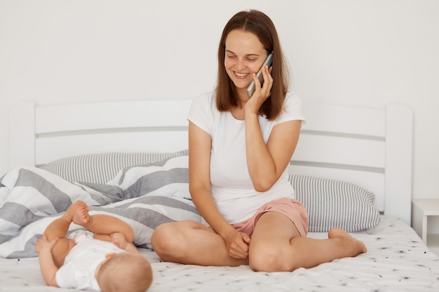 Indoor studio shot of woman wearing white t shirt and shorts talking via smart phone and having positive emotions, looking at infant baby girl, happy motherhood.