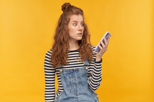 Indoor studio shot of young ginger female with long red curly hair looks aside at phone with shocked facial expression, isolated over yellow background