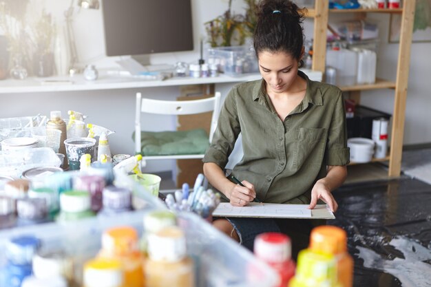 Indoor view of beautiful young Caucasian woman artist with brunette hair busy making drawings in spacious workshop interior with lots of paint bottles