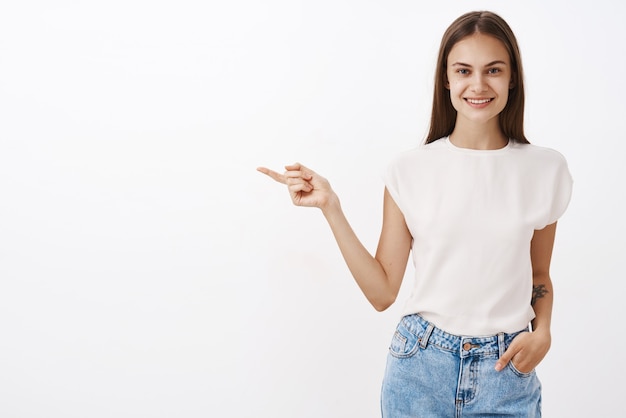 트렌디 한 흰색 티셔츠와 청바지를 입은 매력적인 친근한 여성 동료의 Indoot 샷은 주머니에 친절한 손을 잡고 왼쪽으로 조언을 주거나 방법을 보여줍니다.