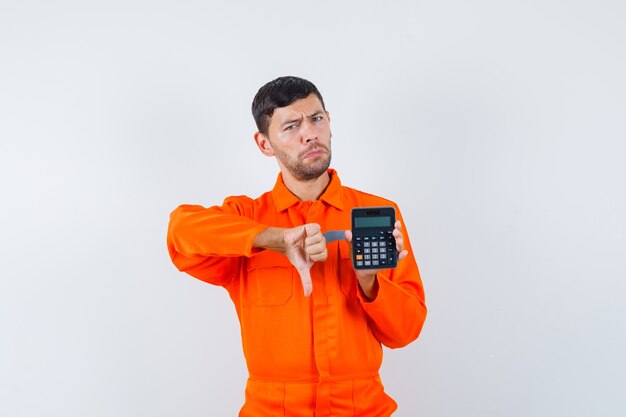 Industrial man holding calculator, showing thumb down in uniform and looking displeased , front view.