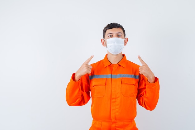 Industrial man pointing at his medical mask in uniform and looking careful , front view.