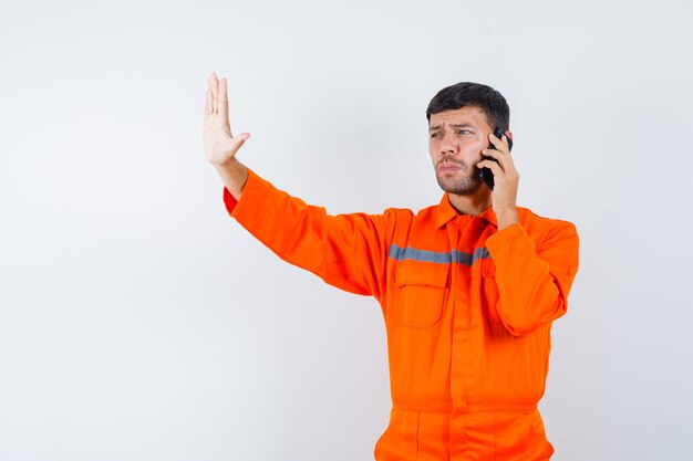Industrial man in uniform talking on mobile phone, showing stop gesture , front view.