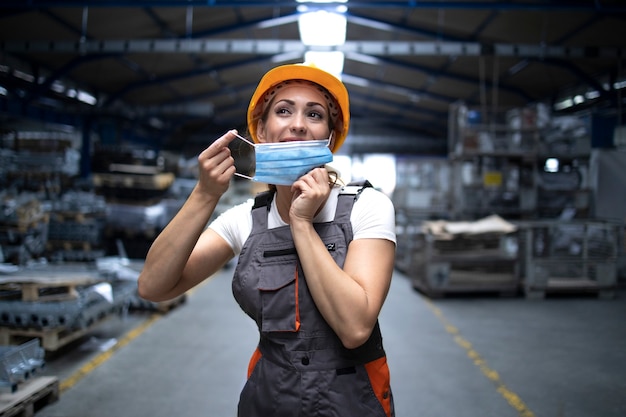 Free photo industrial worker standing in factory hall and putting on hygienic mask on face to protect herself against highly contagious corona virus