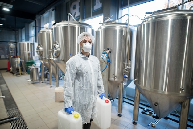 Industrial worker technologist in white protective suit with hairnet and mask holding plastic cans with chemicals in food factory production line