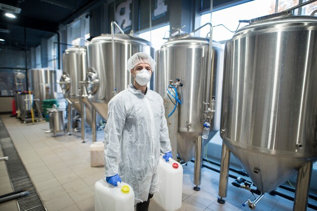 industrial worker technologist in white protective suit with hairnet and mask holding plastic cans with chemicals in food factory production line