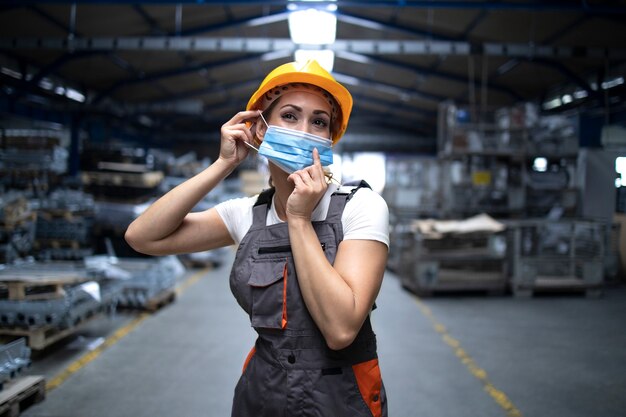 Промышленный рабочий стоит в заводском цехе и надевает гигиеническую маску на лицо, чтобы защитить себя от очень заразного вируса короны