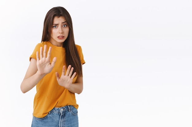 Free photo inescure, reluctant timid scared innocent woman in yellow t-shirt, asking stop or step back, grimacing displeased, rejecting something with awkward, worried expression, pull hands in prohibition