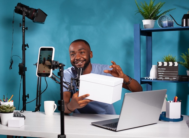 Influencer presenting white mockup gift for fans looking at live video podcast setup in vlogging studio. Smiling vlogger holding product box hosting online giveaway in front of recording smartphone.