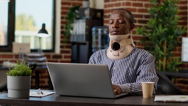 Injured office consultant wearing cervical collar at startup job, using laptop website. Medical neck foam to recover after physical painful vertebrae injury, suffering from spine accident.