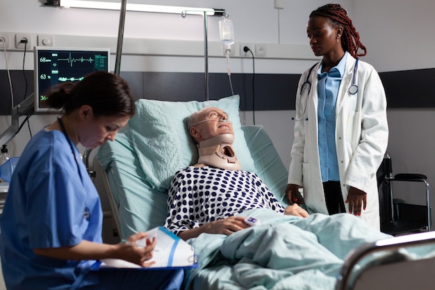 Injured senior man with neck brace laying in bed suffering after accident, discussing with doctor during medical visit and assistant taking notes on clipboard