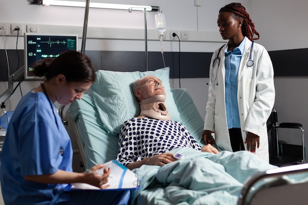 Раненый пожилой мужчина с шейным бандажом, лежащий в постели после несчастного случая, обсуждает с врачом во время посещения врача и помощником, делающим заметки в буфере обмена