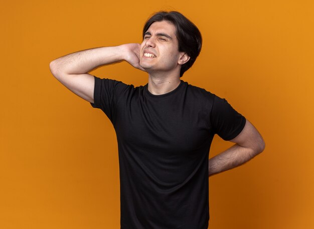 Injured with closed eyes young handsome guy wearing black t-shirt putting hand on aching waist isolated on orange wall