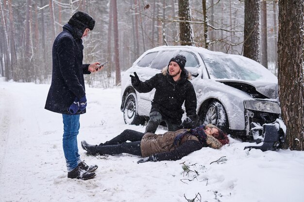 Injured woman is lying on the snow after car crush, man is trying to help her, the second man is calling ambulance.