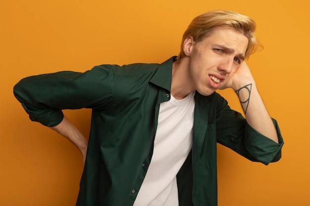 Injured young blonde guy wearing green t-shirt putting hand on aching waist isolated on yellow