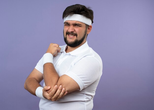 Injured young sporty man wearing headband and wristband grabbed aching elbow isolated on green wall with copy space