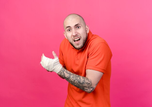 injured young sporty man with wrist bandage grabbed hand isolated on pink