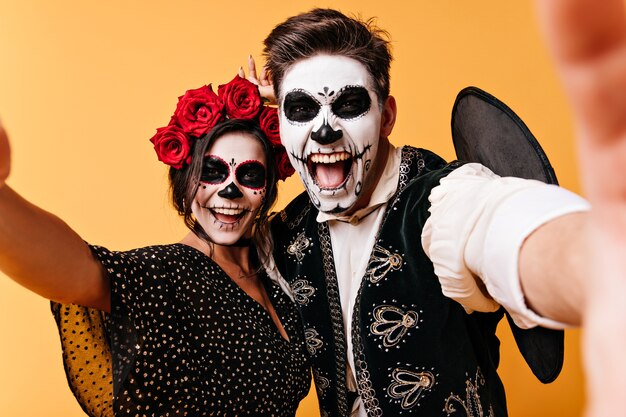 Insane, funny young man and woman take selfies, showing off their skeleton makeup. Girl with flowers on her head and her boyfriend have fun