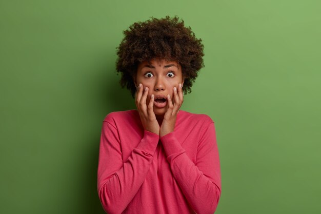 Insecure alarmed African American woman stares anxious , feels shocked and embarrassed, keeps palms on face, wears pink jumper, realizes something shocking isolated on green wall.