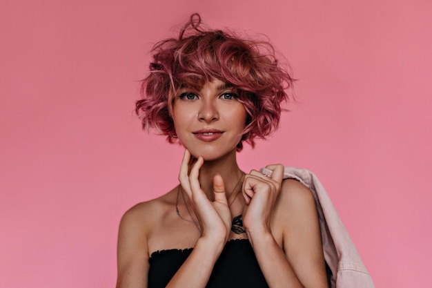 Inside closeup portrait of charming young woman dressed in black cropped top and holding denim pink jacket Cute tanned girl looks into camera and smiles on isolated