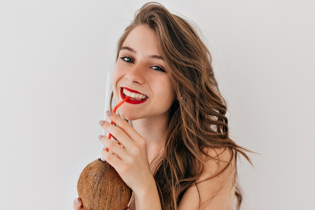 Внутри счастливая стильная женщина с красными губами и белыми зубами и здоровой кожей с вьющимися волосами пьет кокос и позирует