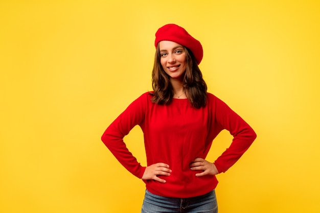 빨간색 세련된 풀오버와 베레모의 짧은 헤어 스타일로 노란색 벽 위에 포즈를 취하는 젊은 유럽 매력적인 아가씨의 내부 사진