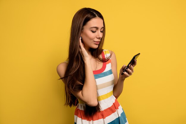 Inside portrait of lovable woman with long hair wearing bright dress holding smartphone and listening music in headphones.