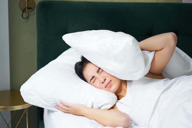 Insomnia asian girl shuts her ears with pillow as partner snoring woman lying in bed bothered by noi