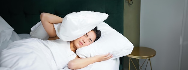 Free photo insomnia asian girl shuts her ears with pillow as partner snoring woman lying in bed bothered by