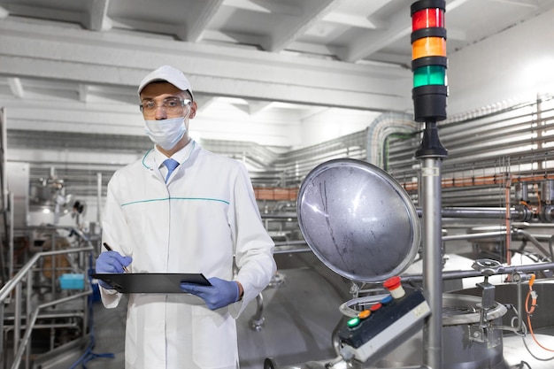 Inspector in a mask and a scrub stands with a foldertablet in his hands at the dairy plant