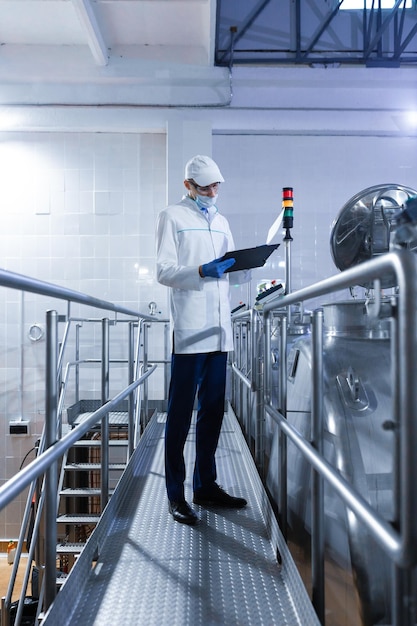 Inspector in a mask and a scrub stands with a foldertablet in his hands at the dairy plant