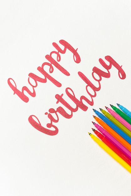 Inspirational phrase 'Happy birthday' for greeting cards and posters drawing with red marker on white paper. Top view of lettering, bunch of colourful markers